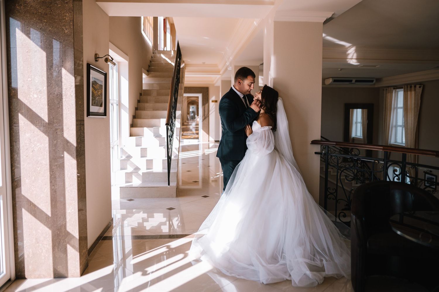 A stunning wedding dress is displayed in a sunlit hallway with elegant architectural details and warm lighting.
