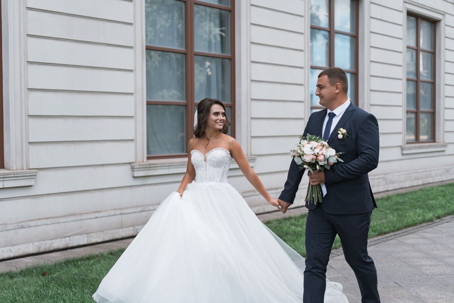 A bride in a white ballgown and groom in dark suit walk together outside a classic stone building.