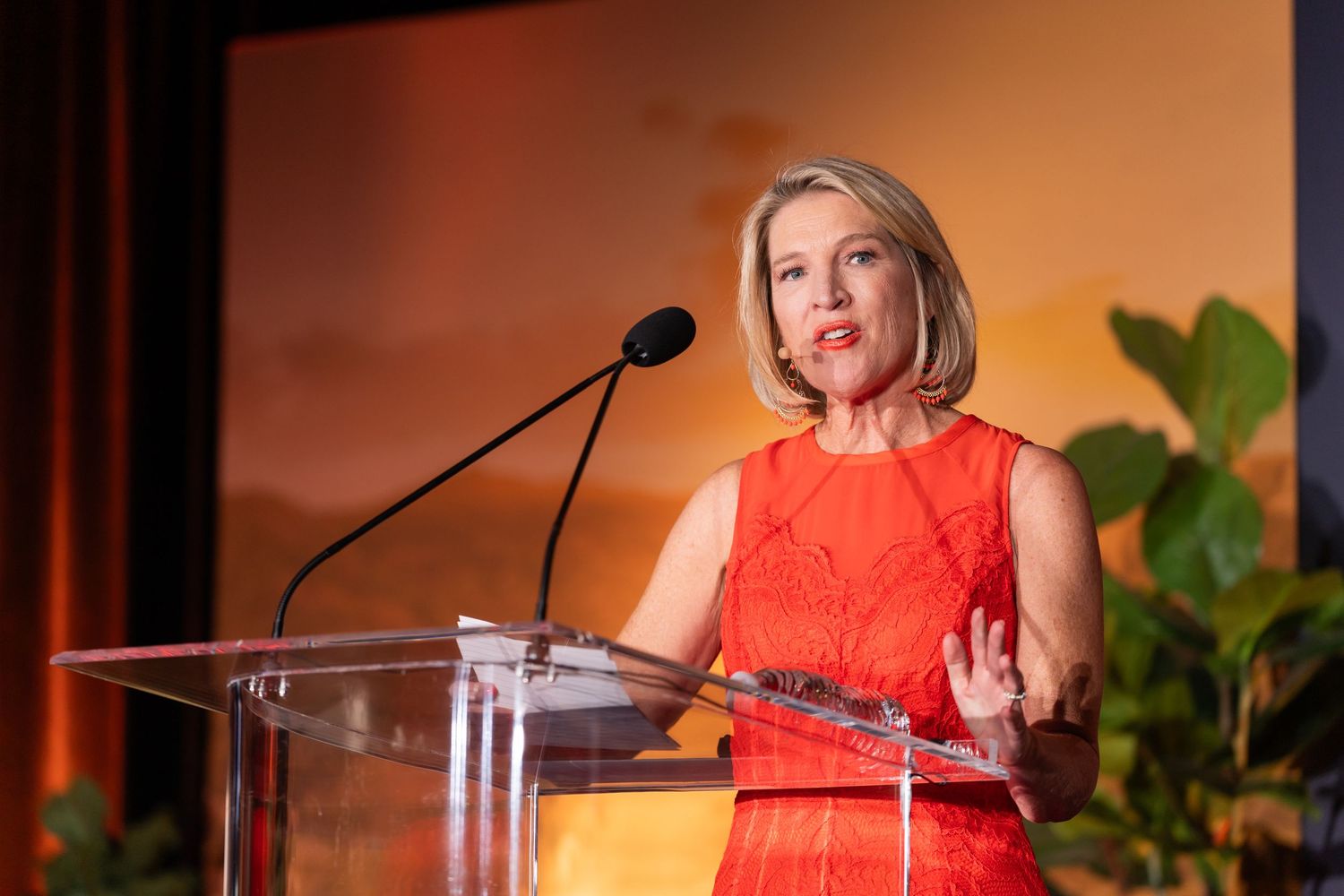 Speaker in bright orange dress delivers remarks at podium during formal event.