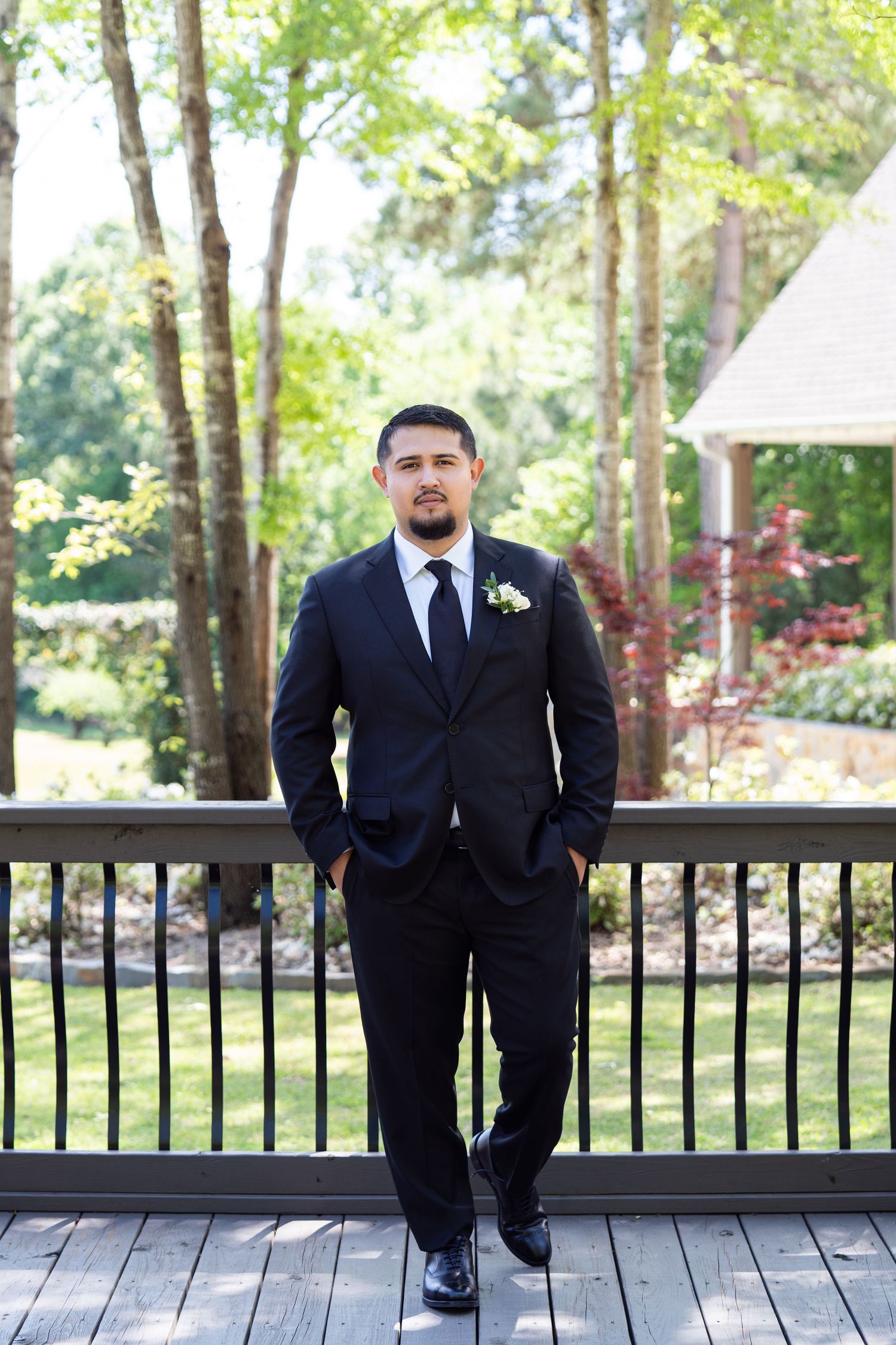 Groomsmen in black suits stand on wooden deck railing at outdoor wedding venue surrounded by trees.