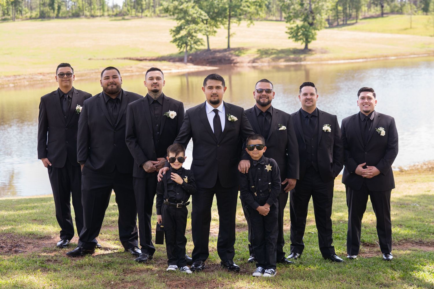 A wedding party in matching black suits poses together by a scenic lake on a sunny day.