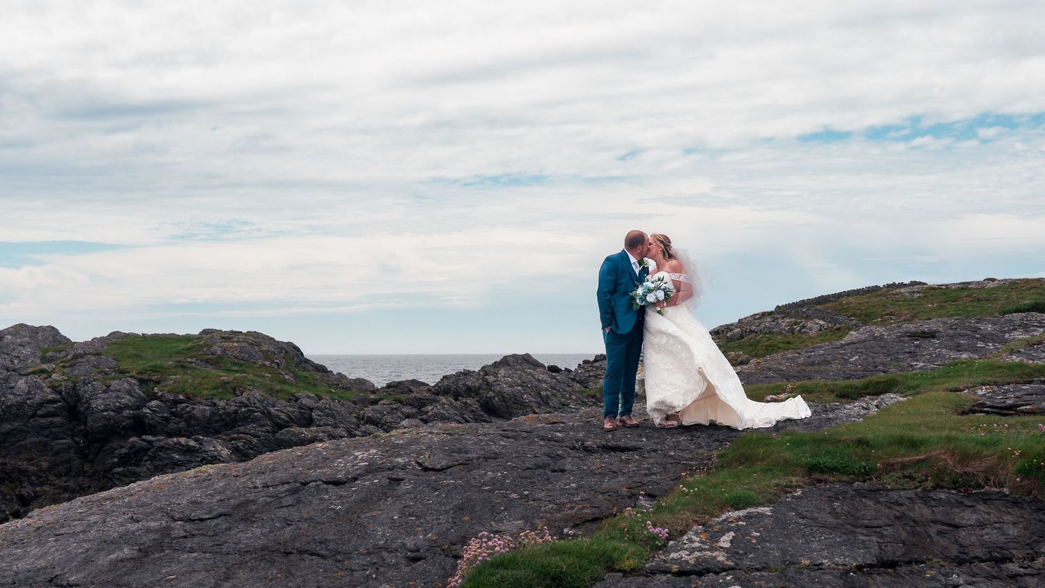 A bride and groom share a romantic kiss on rocky coastal cliffs with the ocean and cloudy sky in the background.