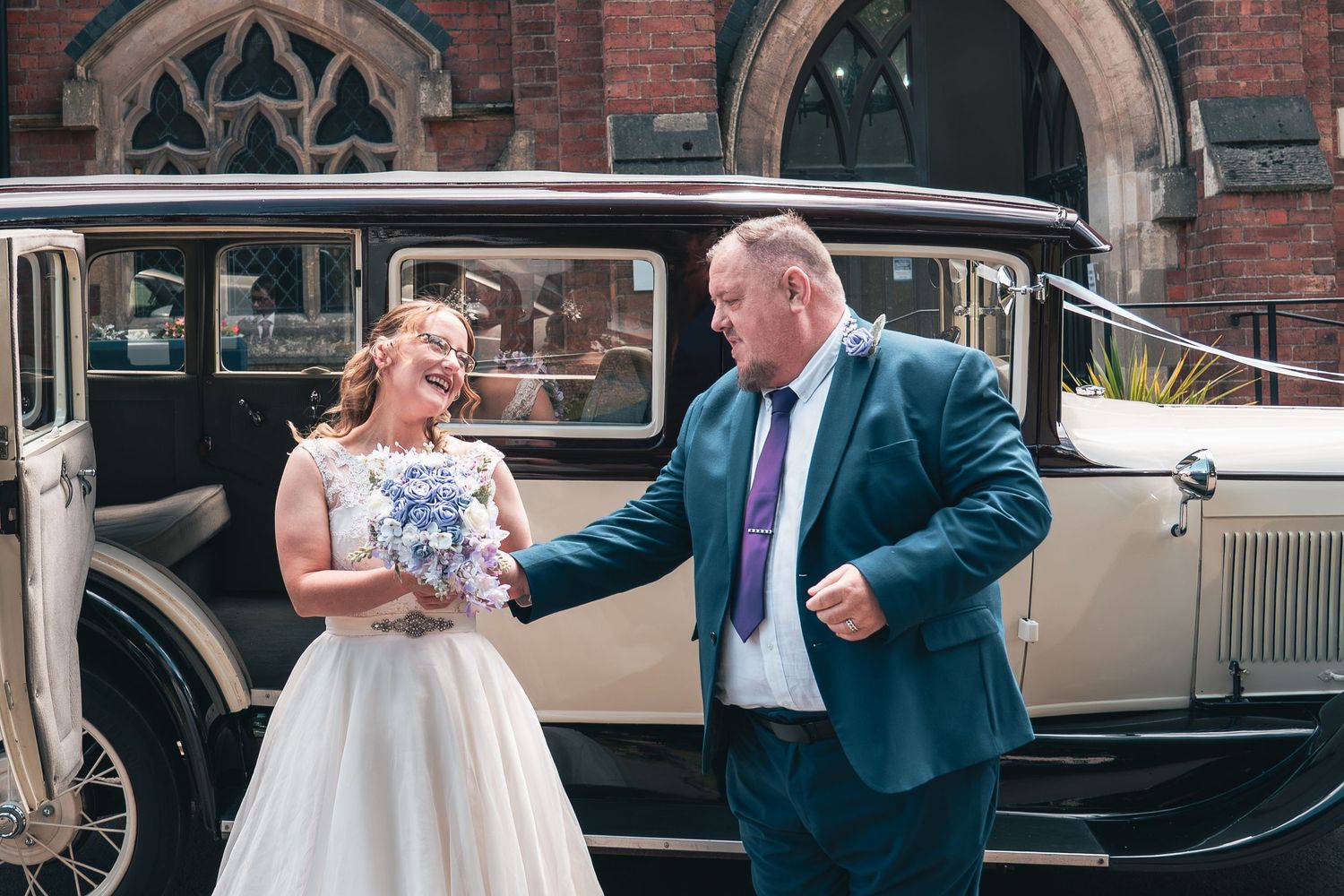 Bride and groom smiling beside a vintage cream wedding car outside a historic brick church.
