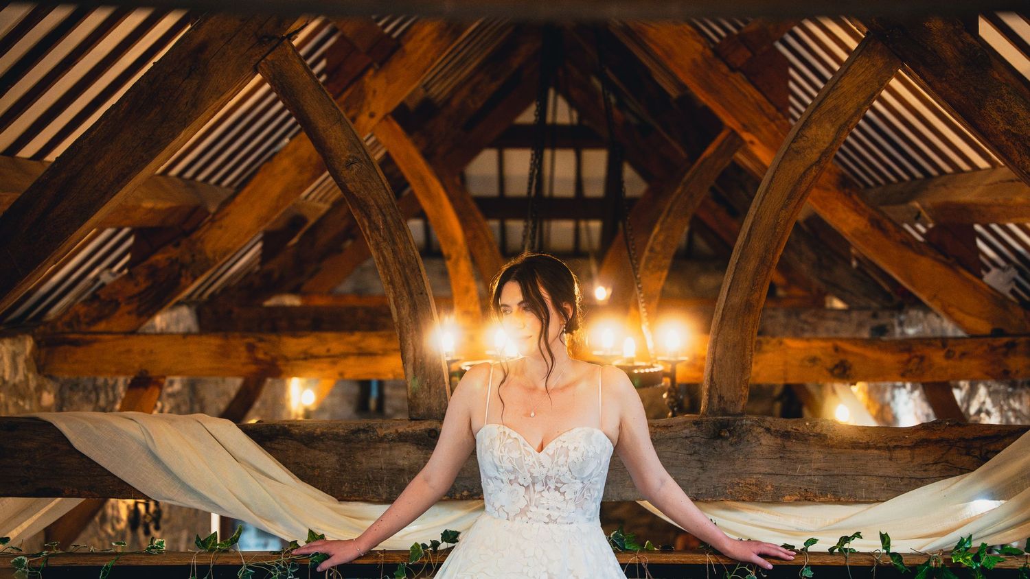 A bride in a white lace dress stands gracefully in a rustic barn venue with exposed wooden beams above.