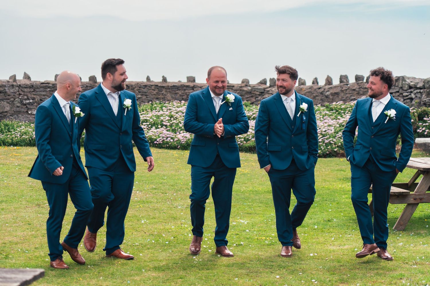Five groomsmen in matching teal suits laughing and walking together outdoors at a wedding venue with stone walls.