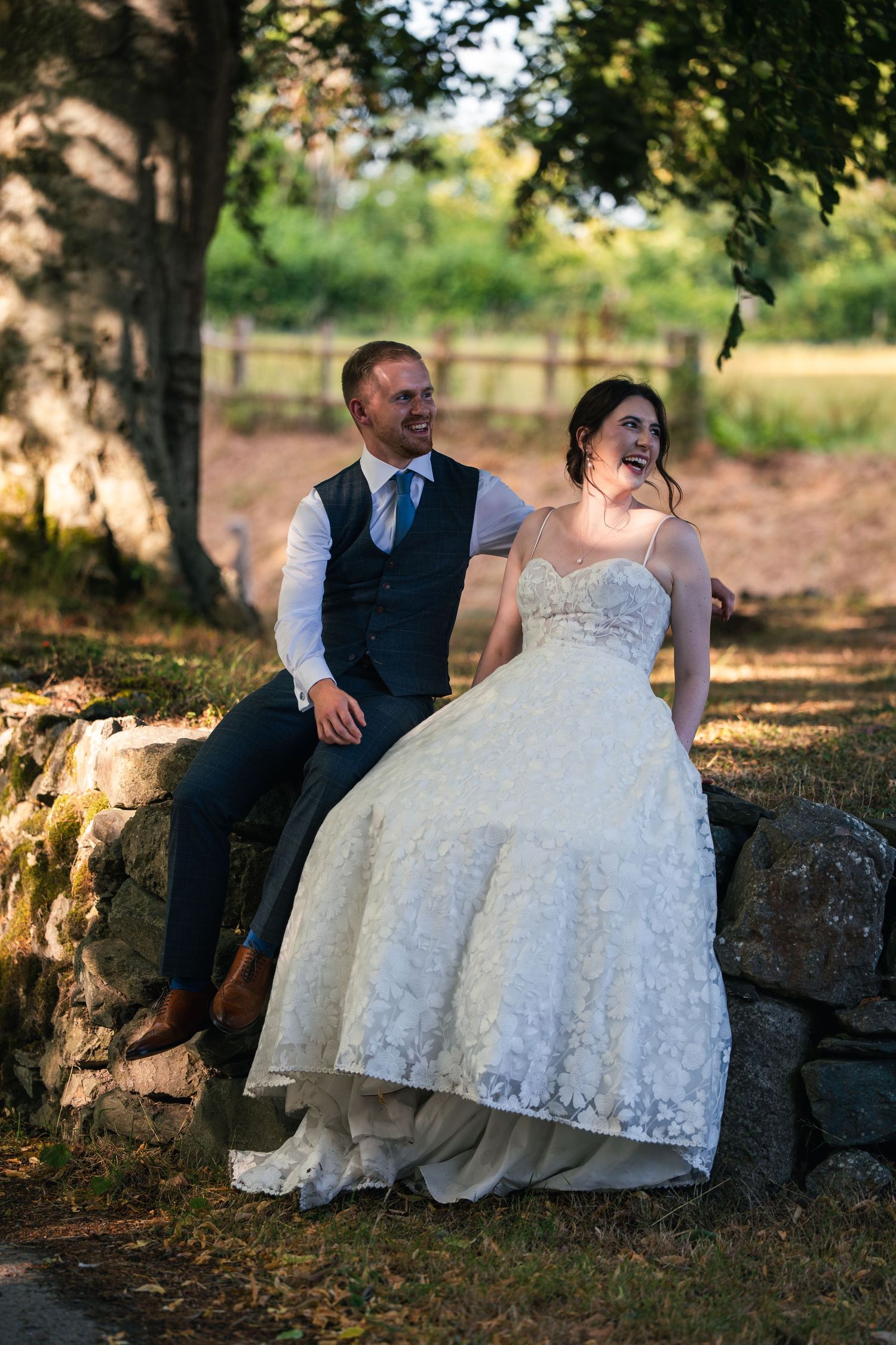 A smiling bride in a white lace gown and groom in a navy vest sit together on a stone wall outdoors.
