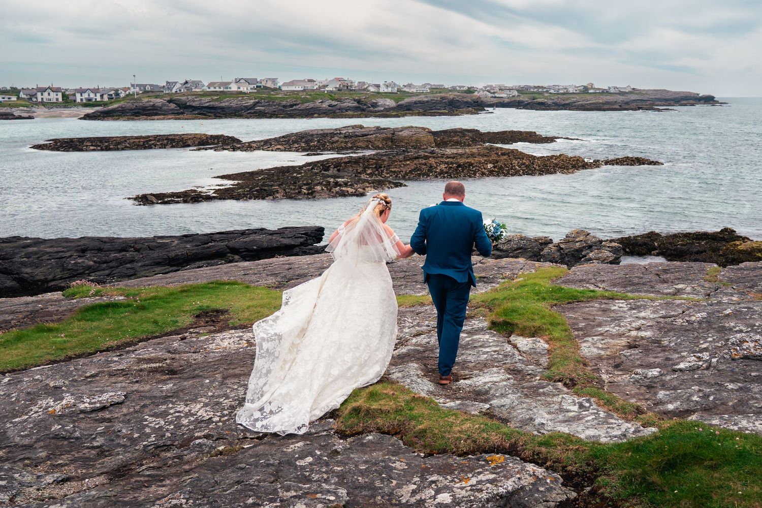 Bride in lace gown and groom in teal suit walking hand in hand across rocky Irish coastal landscape.
