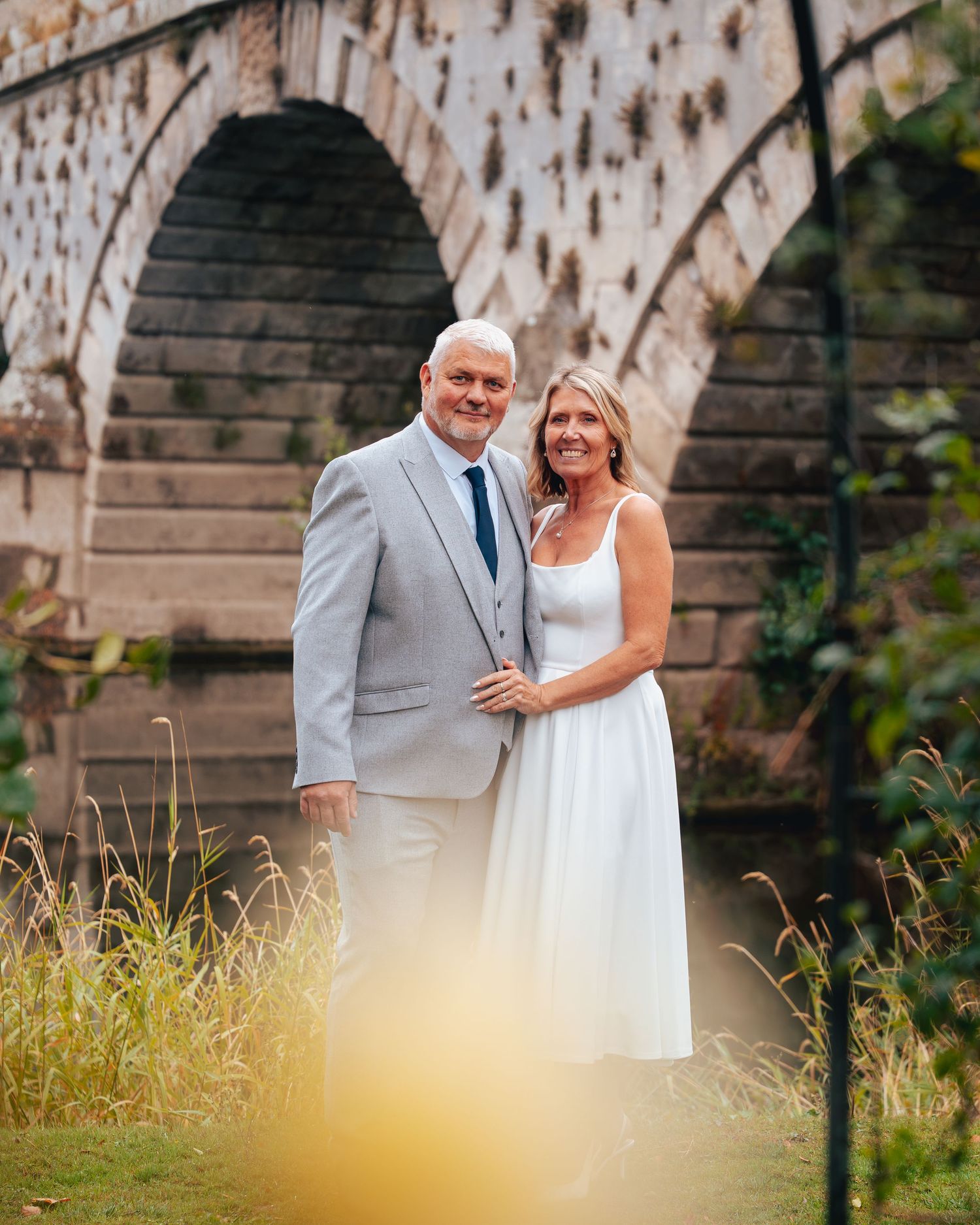 Elegant mature couple in wedding attire posing before a stone arch bridge surrounded by golden grass.