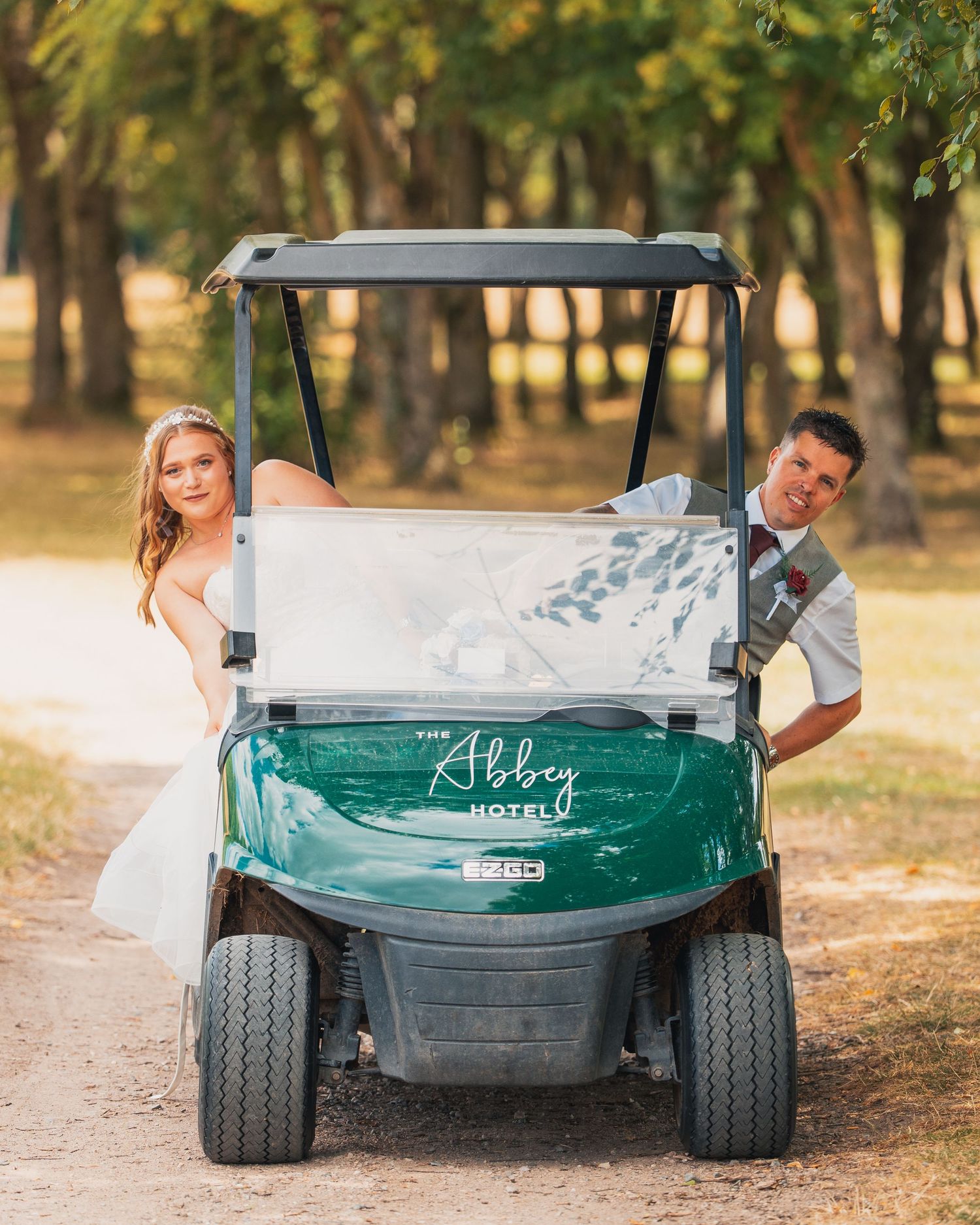 A bride and groom pose playfully with a green Abbey Hotel golf cart on a tree-lined path on their wedding day.