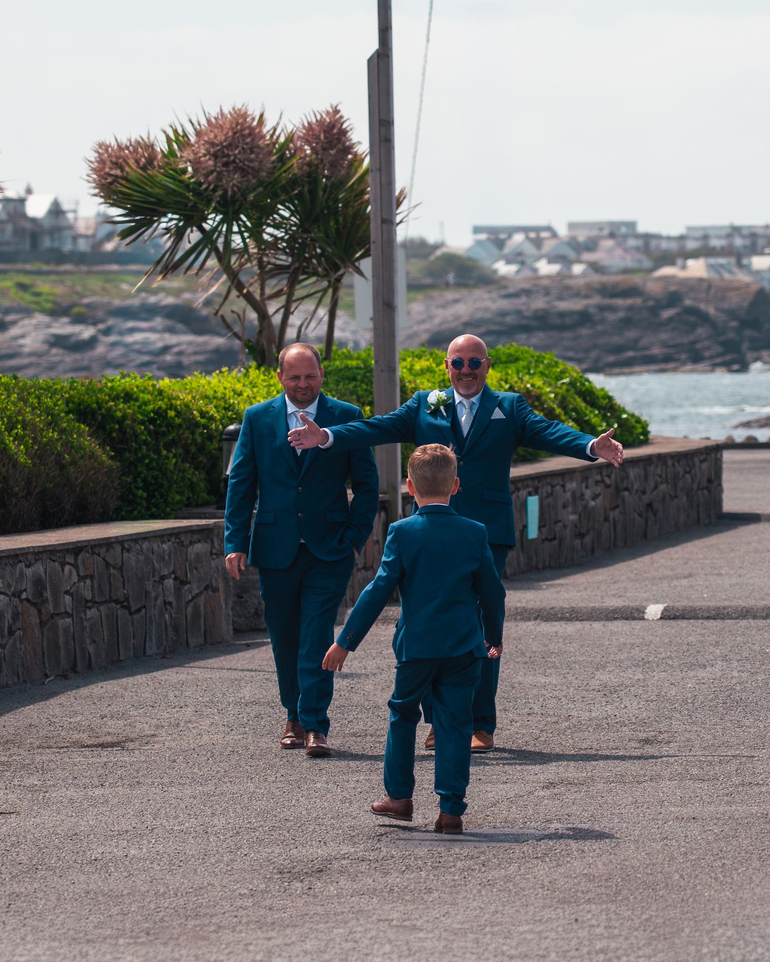 Groom in teal suit welcomes young boy running toward him on coastal wedding day path.