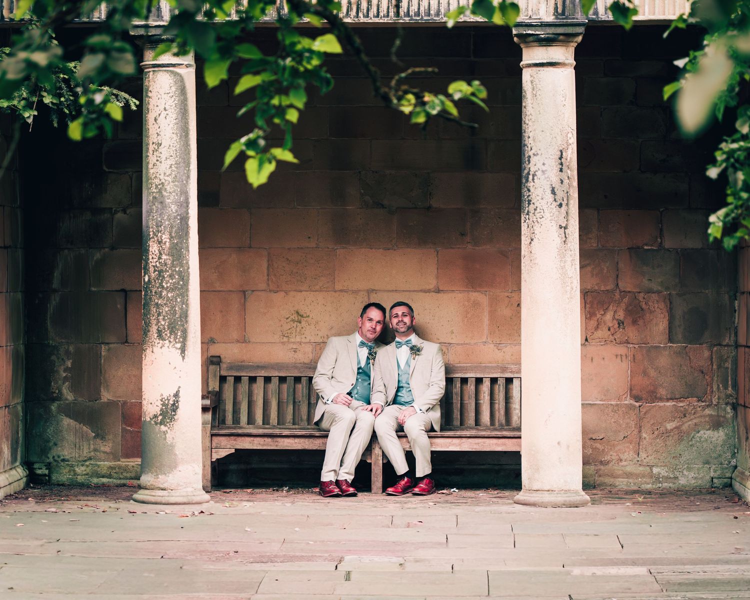 Two grooms in matching suits sit closely on a stone bench between classical columns in a historic garden setting.