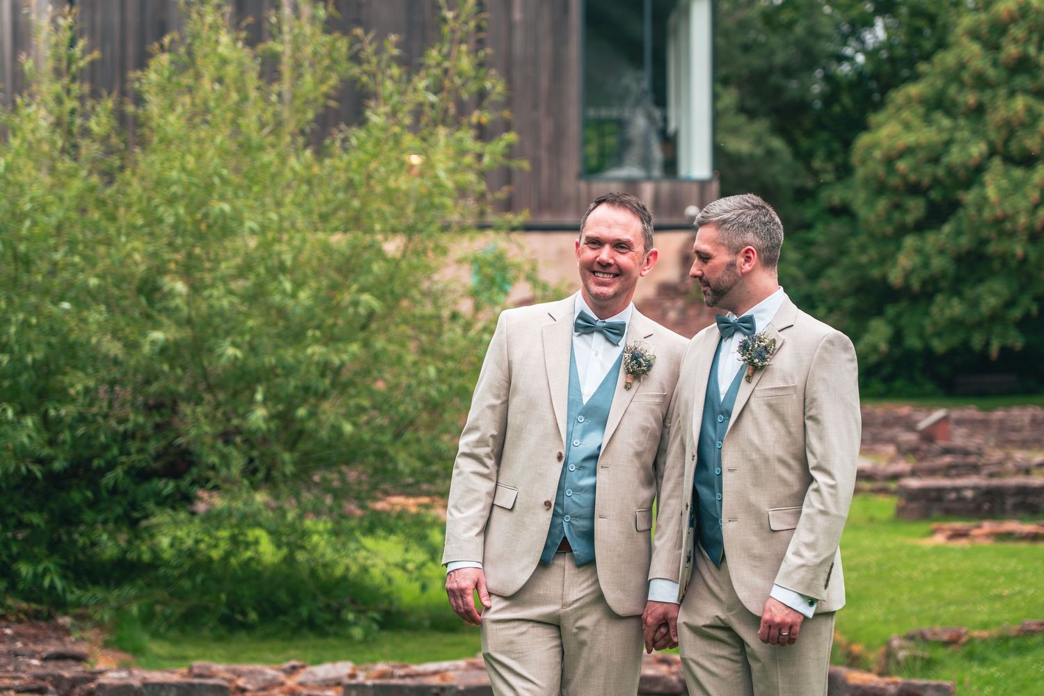 Two grooms in beige suits with teal vests smile and laugh together outdoors.
