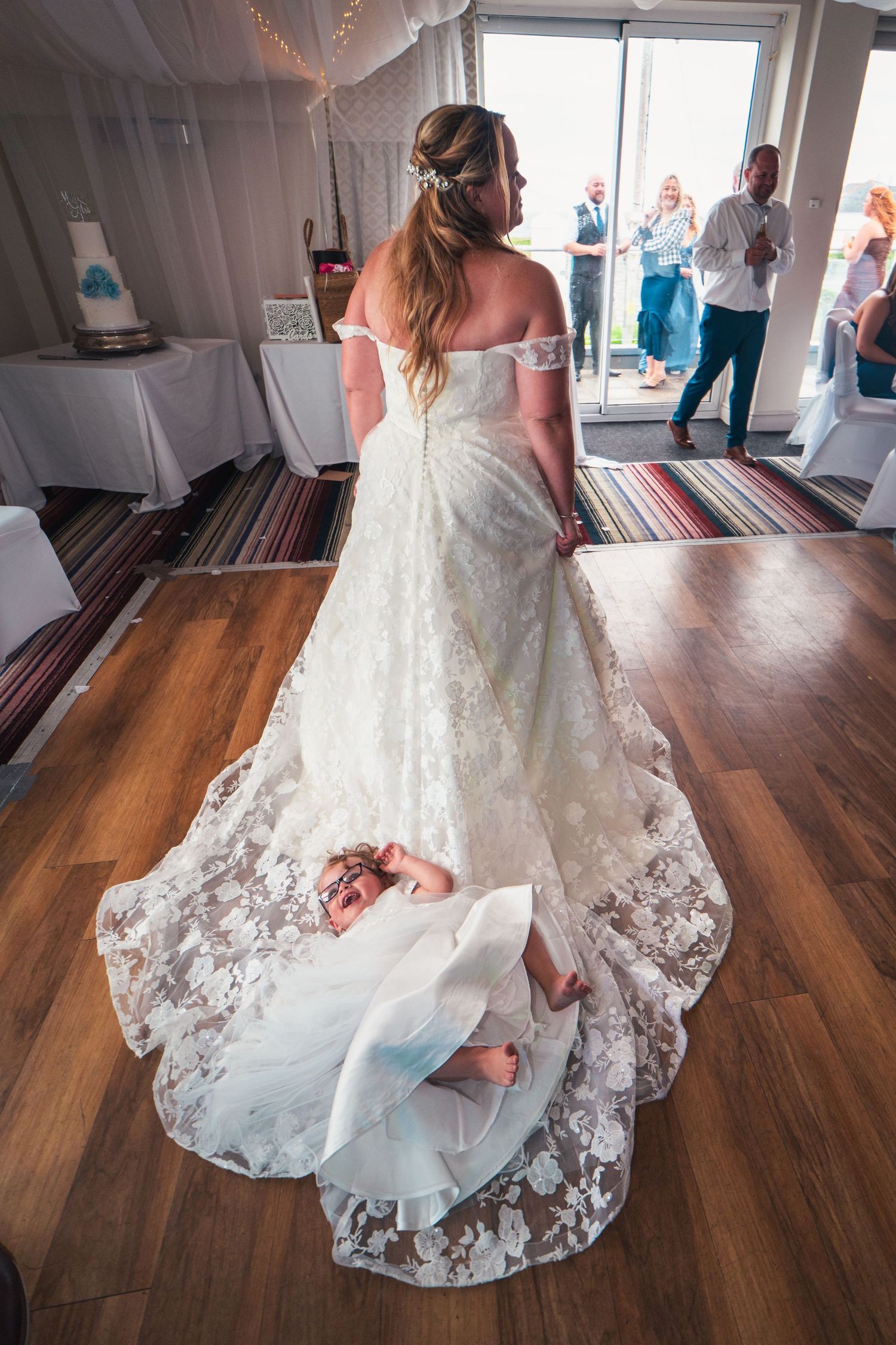 Bride in off-shoulder lace wedding gown with long train on dance floor, child hiding underneath.