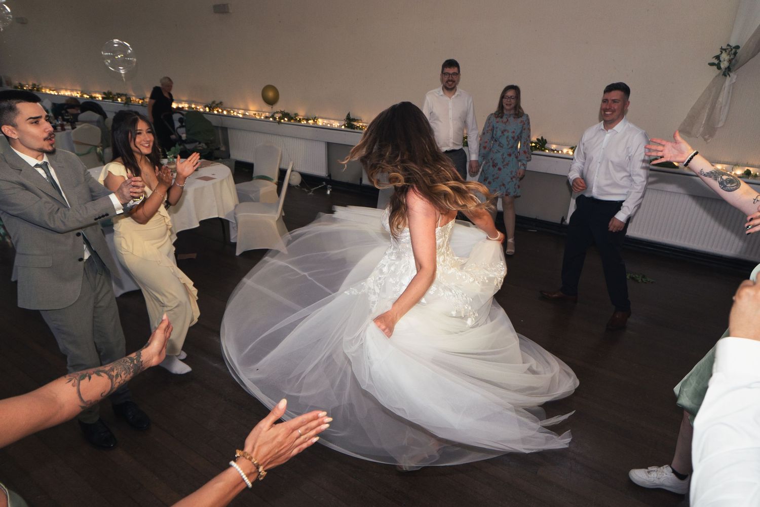 Bride twirling in white wedding gown on dance floor surrounded by guests at elegant indoor reception.