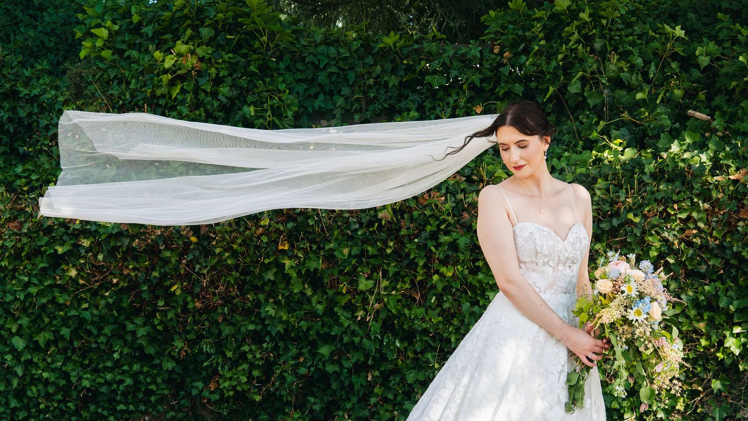 A bride in a white lace gown smiles softly as her flowing veil billows in the breeze against green foliage.