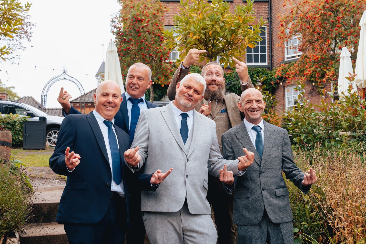 Five men in suits celebrate joyfully in an autumn garden outside a brick wedding venue.