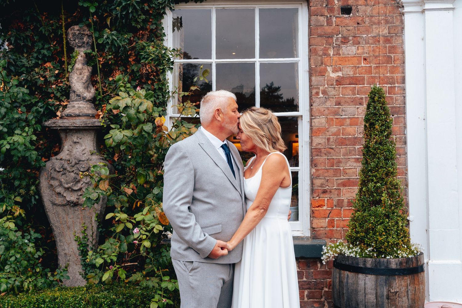 A couple shares a romantic kiss outside a brick building, dressed in wedding attire, surrounded by lush greenery.