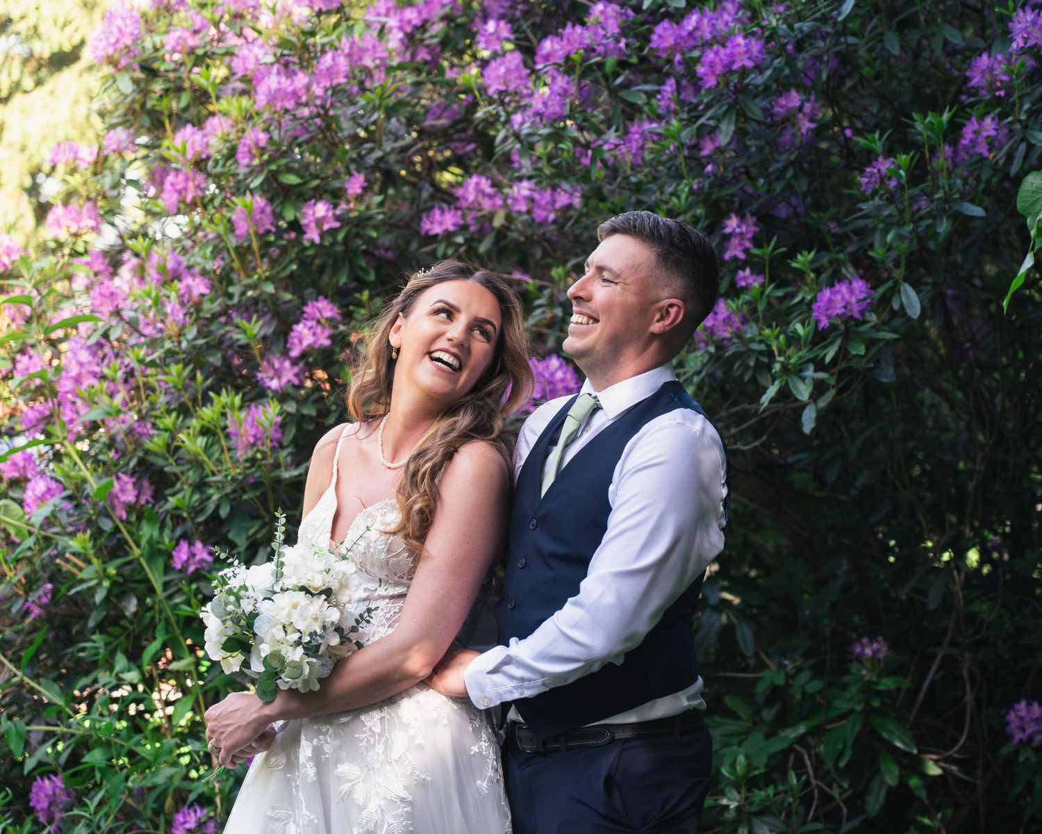 Smiling bride and groom embracing in front of vibrant purple flowering bushes outdoors.