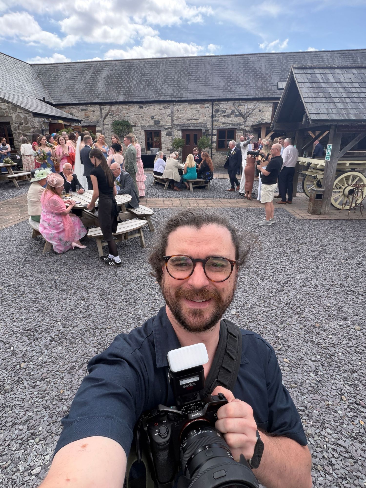 Photographer taking a selfie with a camera at an outdoor wedding reception venue with guests socializing in background.