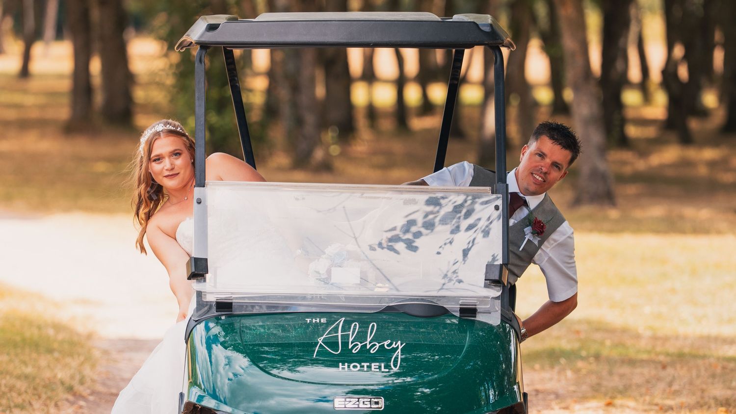 Bride and groom riding a green Abbey Hotel golf cart outdoors, surrounded by trees on their wedding day.