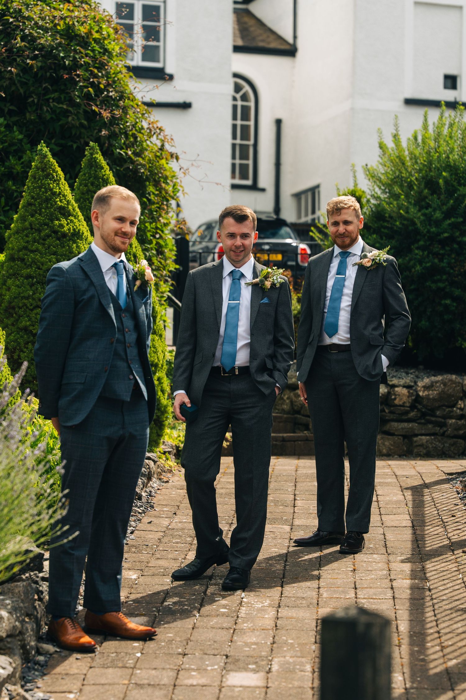 Three groomsmen in dark suits with blue ties and boutonnieres standing outside a white building on a brick path.