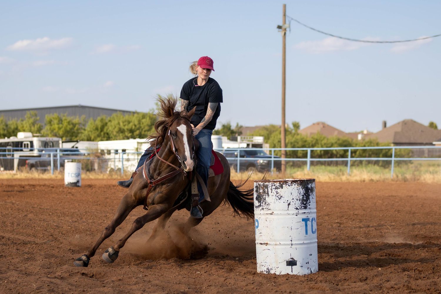 Female rider in pink cap racing horse around barrel in dusty outdoor rodeo arena.