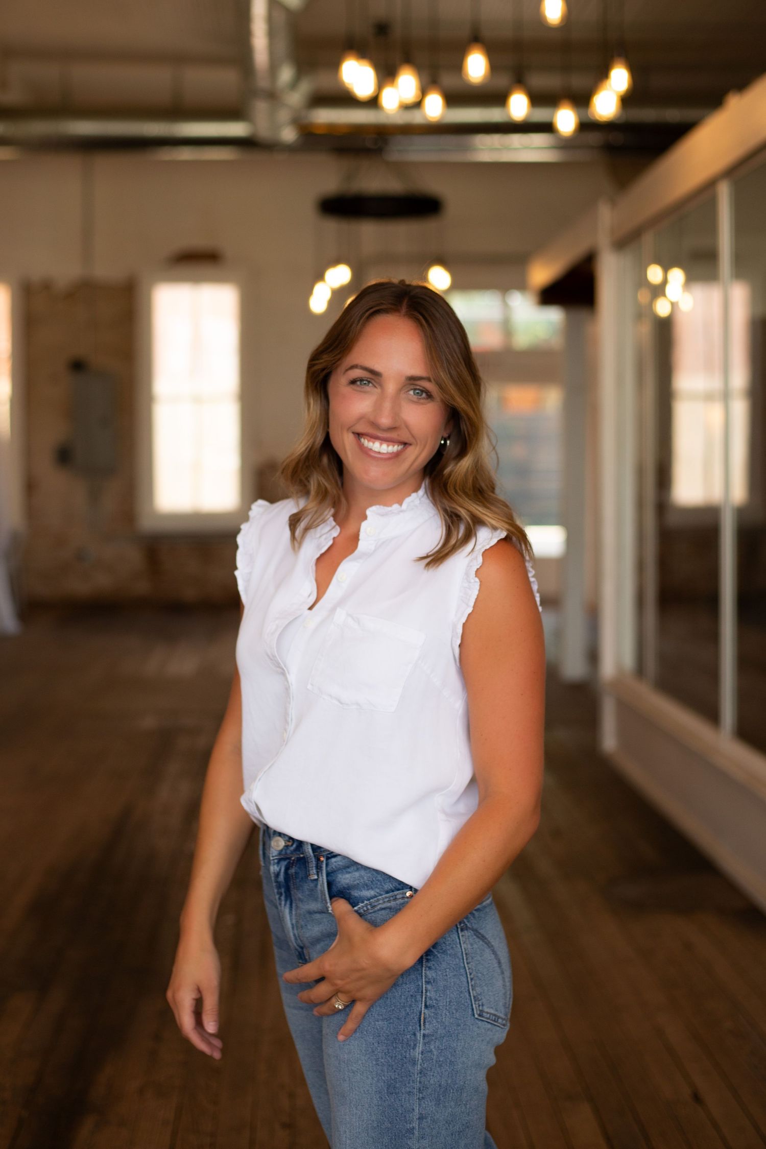 Smiling woman in white sleeveless top and jeans posing in a warmly lit rustic indoor space.