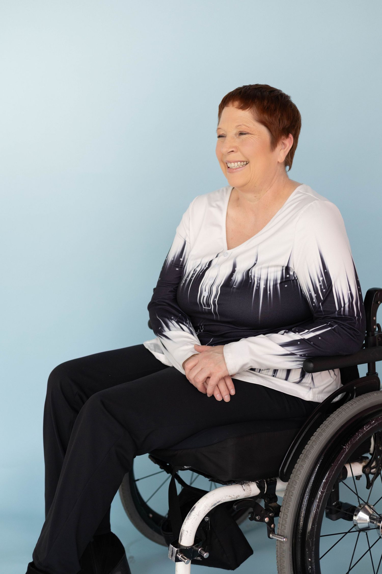 Smiling middle-aged woman with short red hair sitting in wheelchair against light blue background.