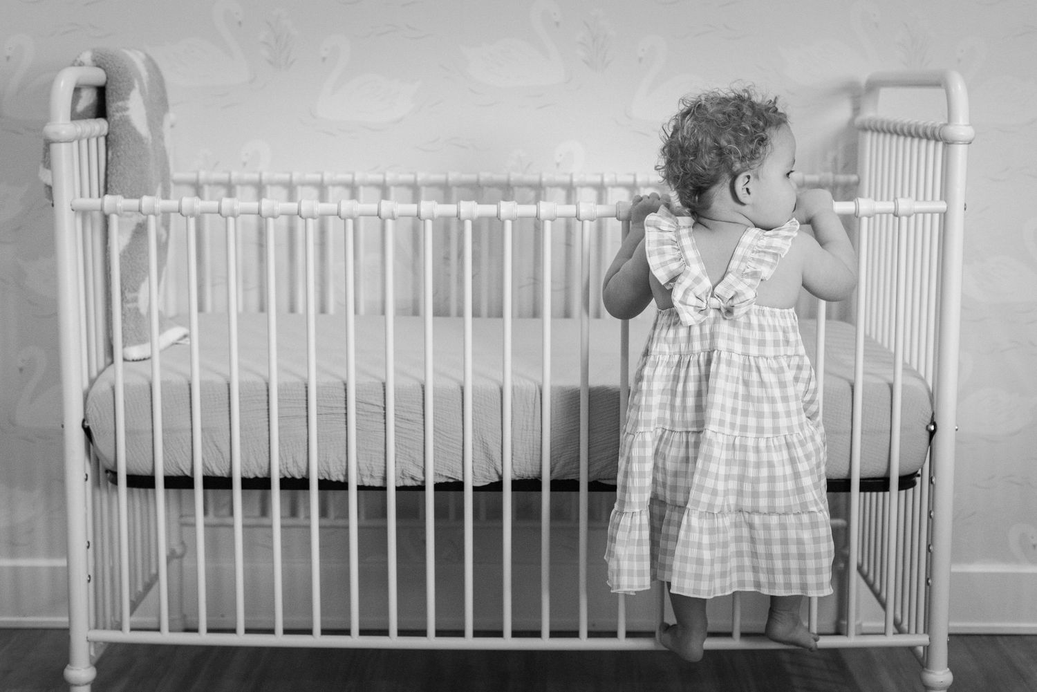 Toddler in a plaid dress standing beside a white vintage iron crib in a nursery, black and white photo.