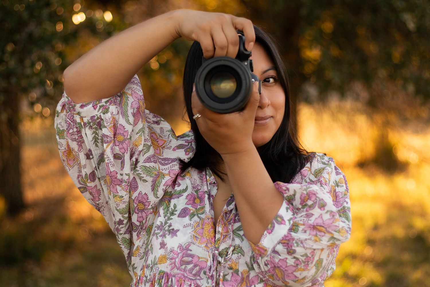 Woman in floral dress photographing outdoors with professional DSLR camera in golden hour light.