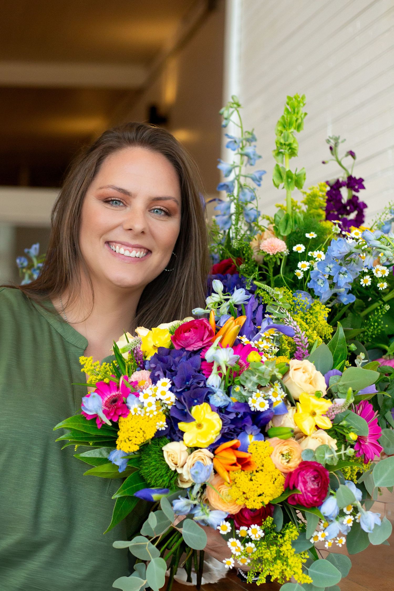 Smiling brunette woman with green eyes holding a large vibrant mixed flower bouquet outdoors.