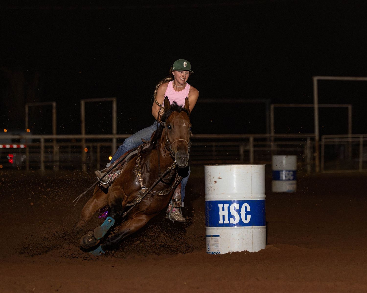A female barrel racer on a brown horse rounds an HSC barrel at a night rodeo competition.