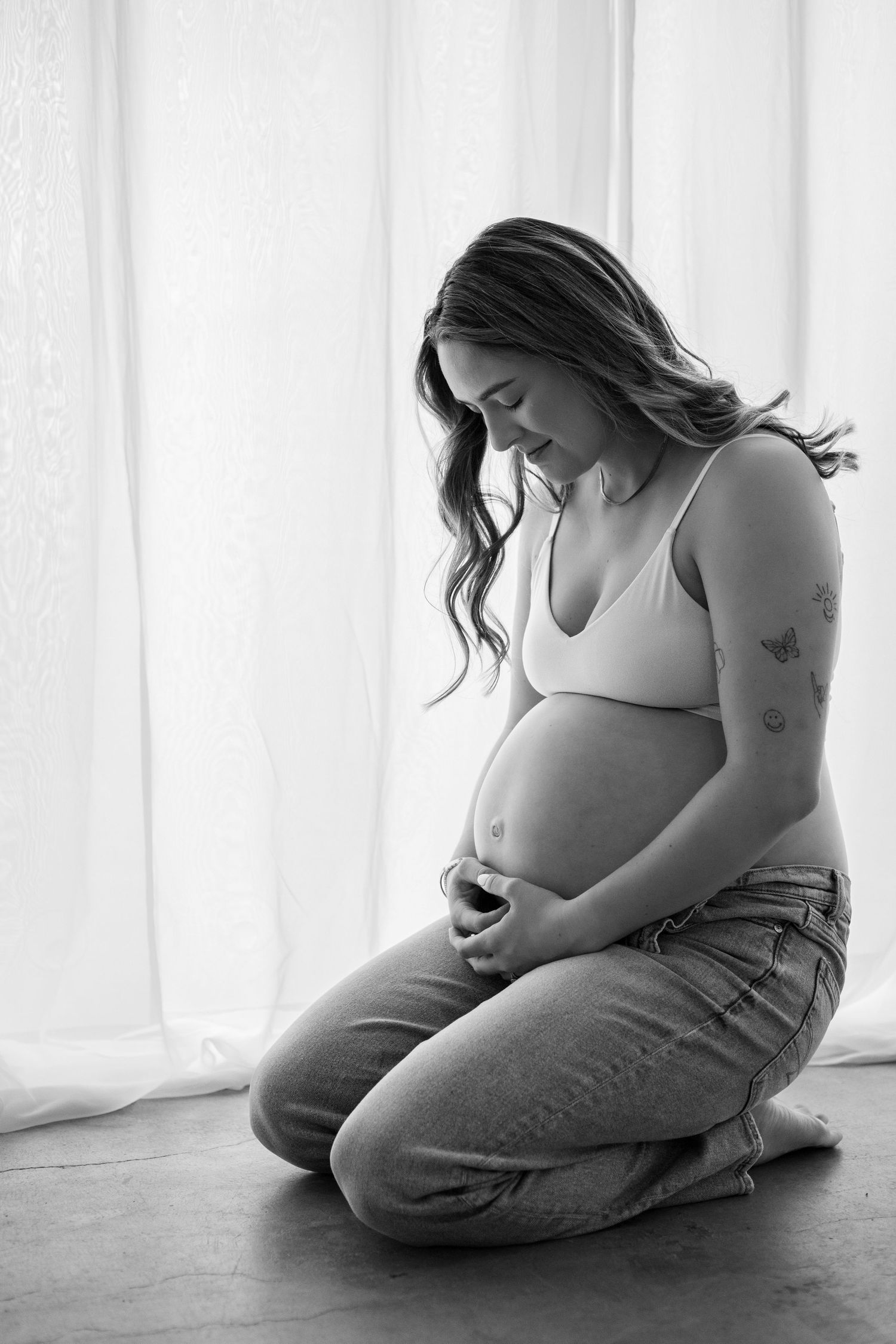 Black and white maternity photo of pregnant woman kneeling, cradling baby bump, wearing jeans and bralette near sheer curtains.