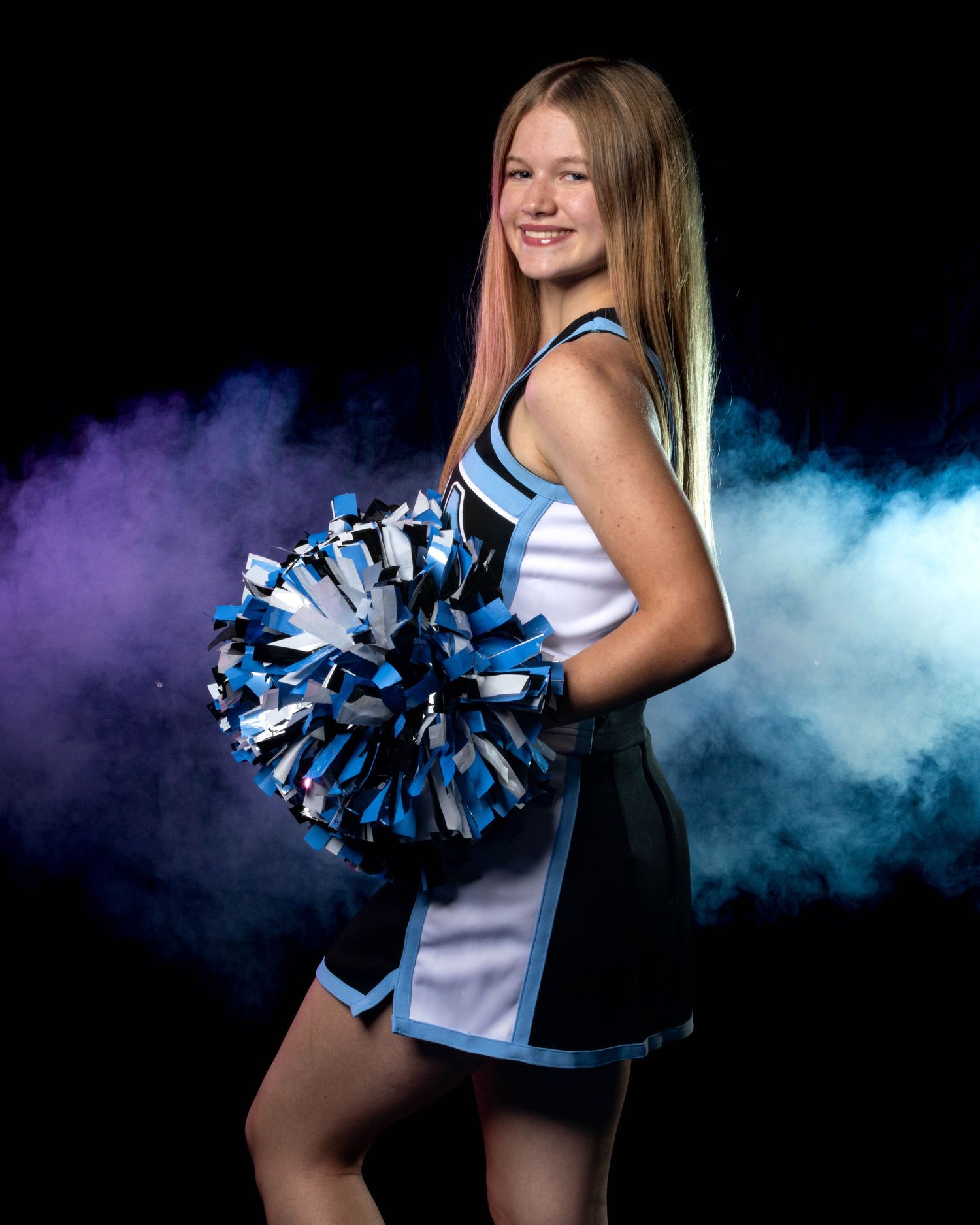 Smiling cheerleader in blue and white uniform holds silver and blue pom-poms in a smoky studio portrait.