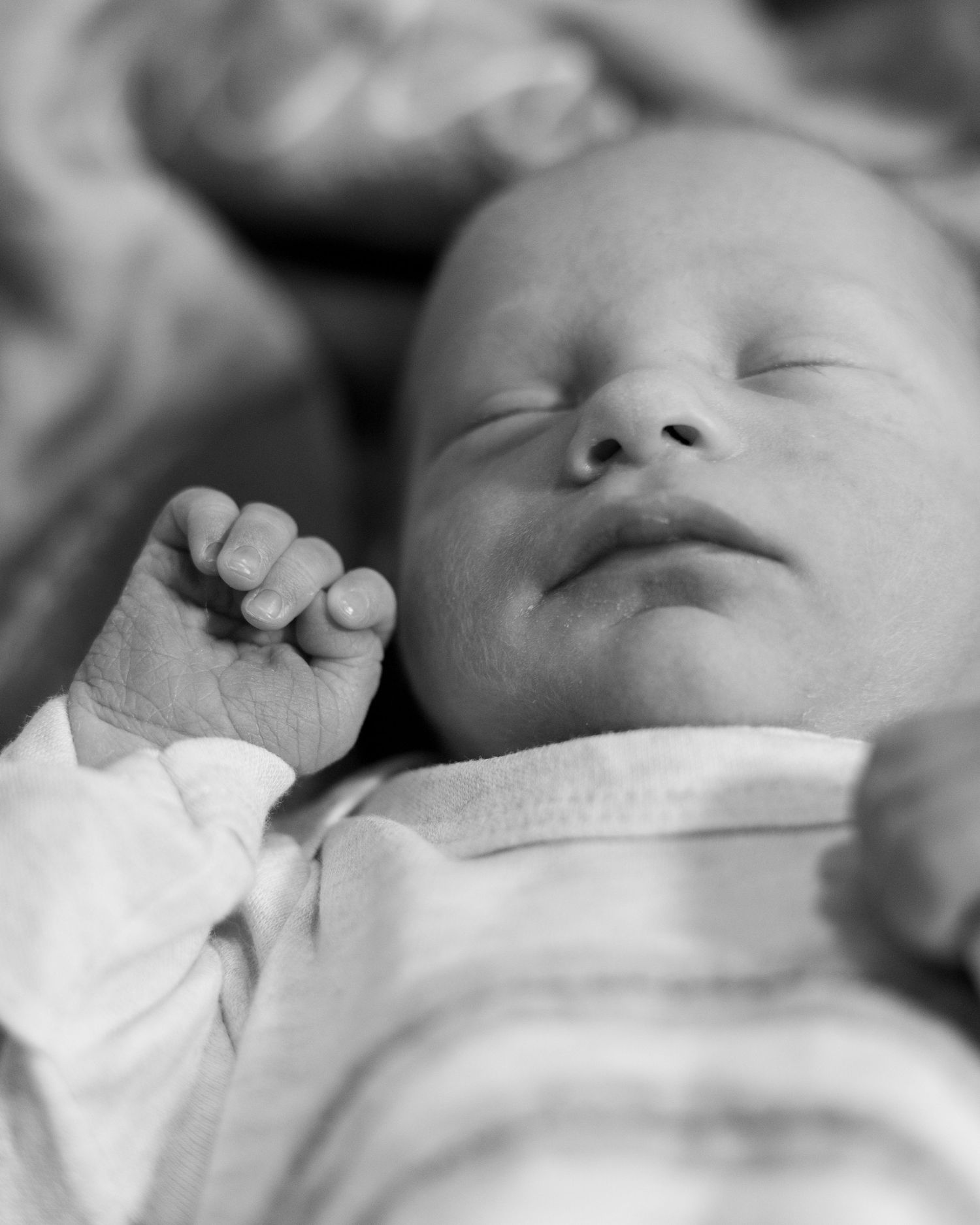 Black and white photo of a peaceful sleeping newborn baby with tiny clenched fist, wearing a striped onesie.