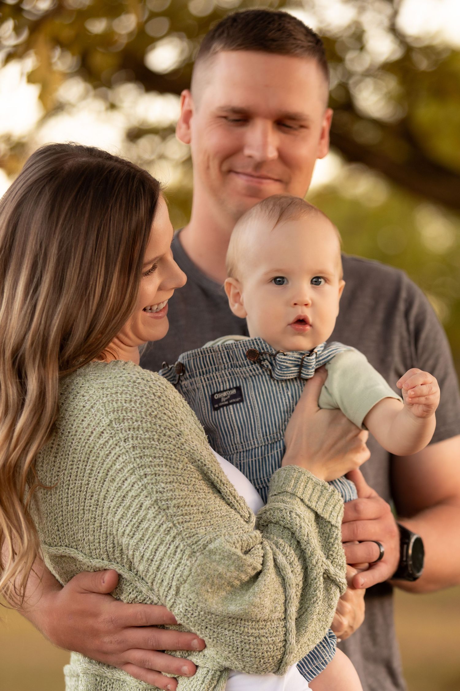 Smiling parents holding adorable baby boy outdoors during golden hour.