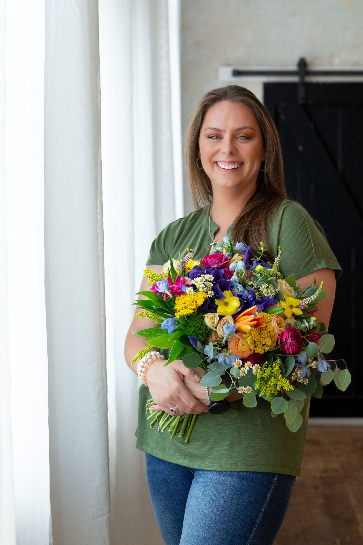Smiling brunette woman in green shirt holding a vibrant colorful flower bouquet indoors.