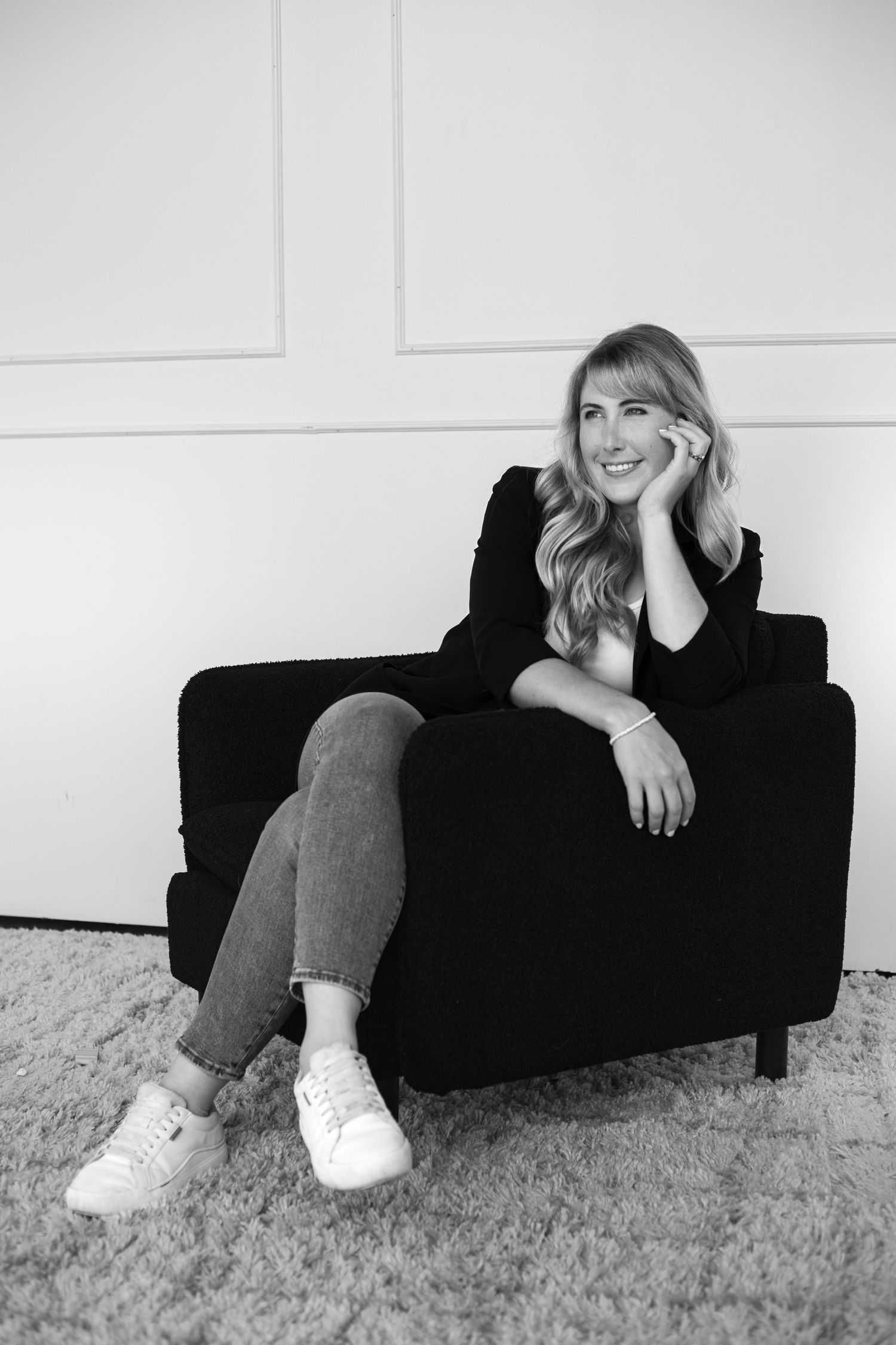 Cheerful blonde woman in black blazer relaxing in armchair, black and white portrait photo.