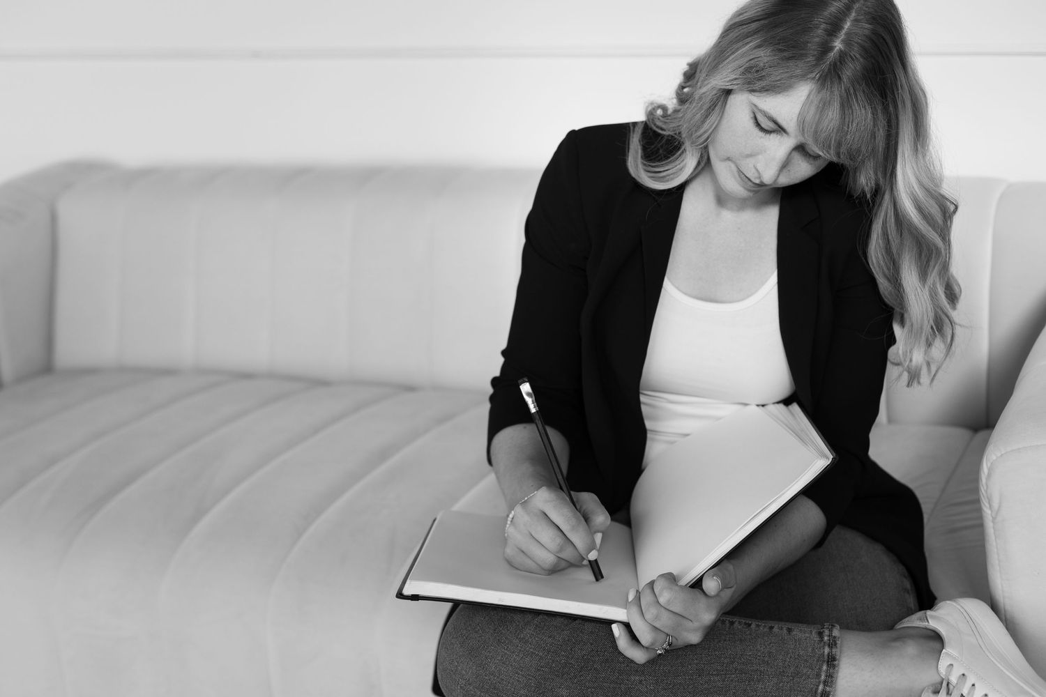 Black and white image of blonde woman writing in notebook while seated on couch.