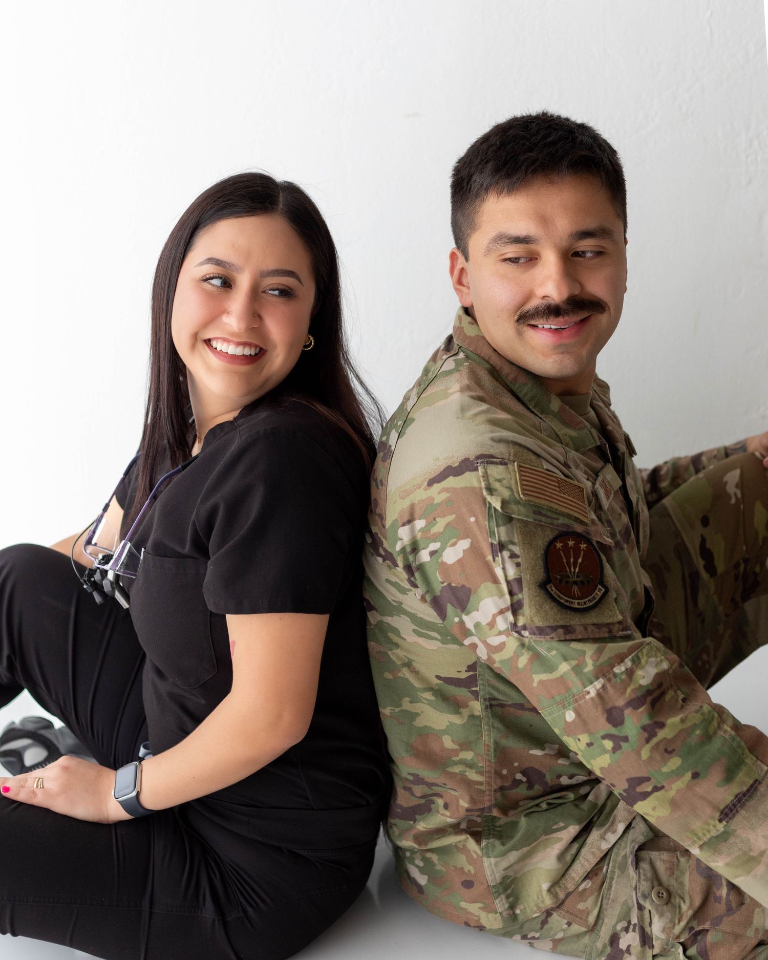 Smiling couple sitting back to back, woman in black scrubs and man in military camouflage uniform.