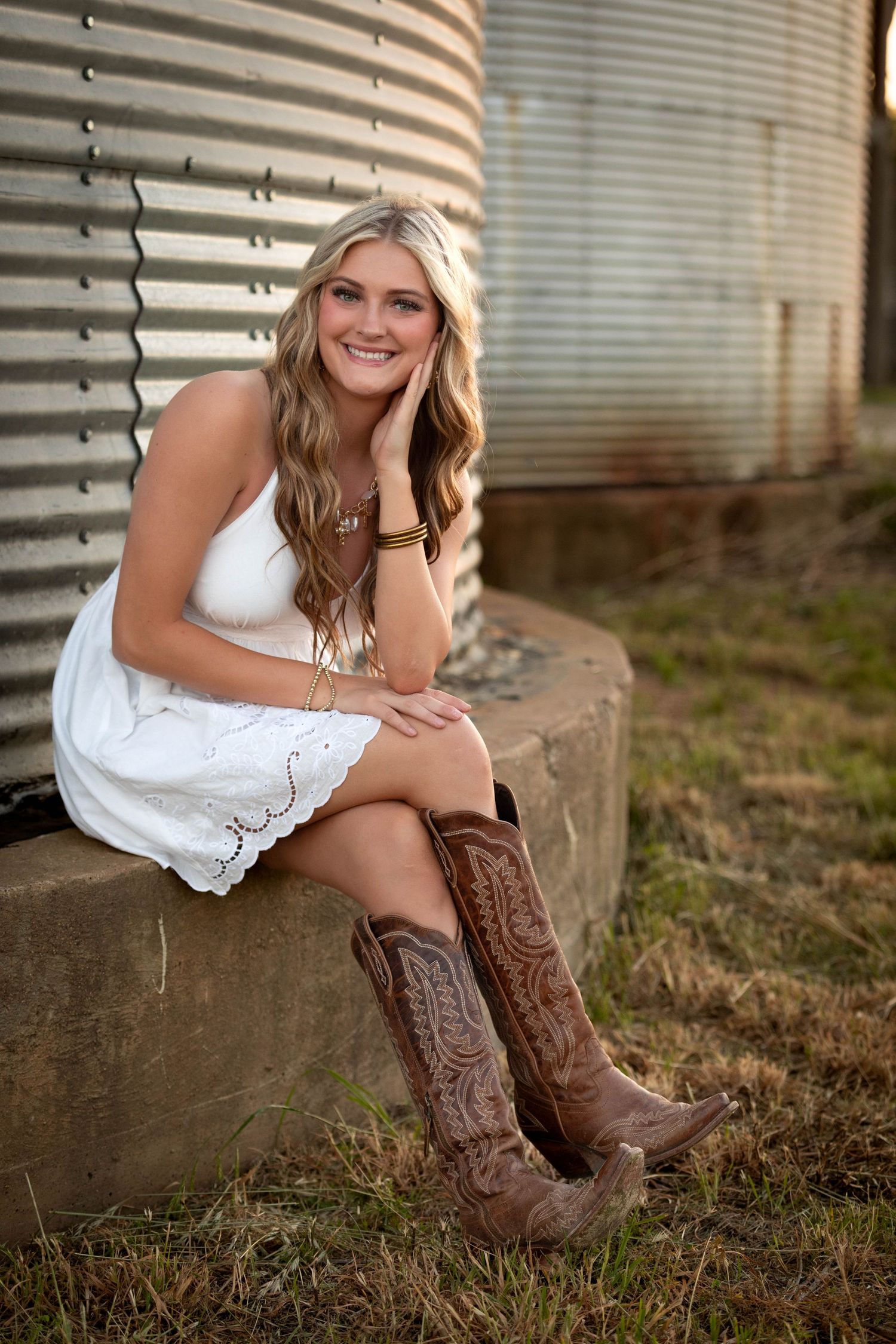 Smiling blonde teen in white sundress and brown cowboy boots seated near a rustic grain silo.