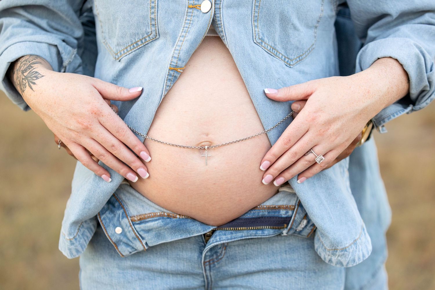 Pregnant woman in denim outfit holding baby bump, showing belly button piercing and tattoo outdoors.