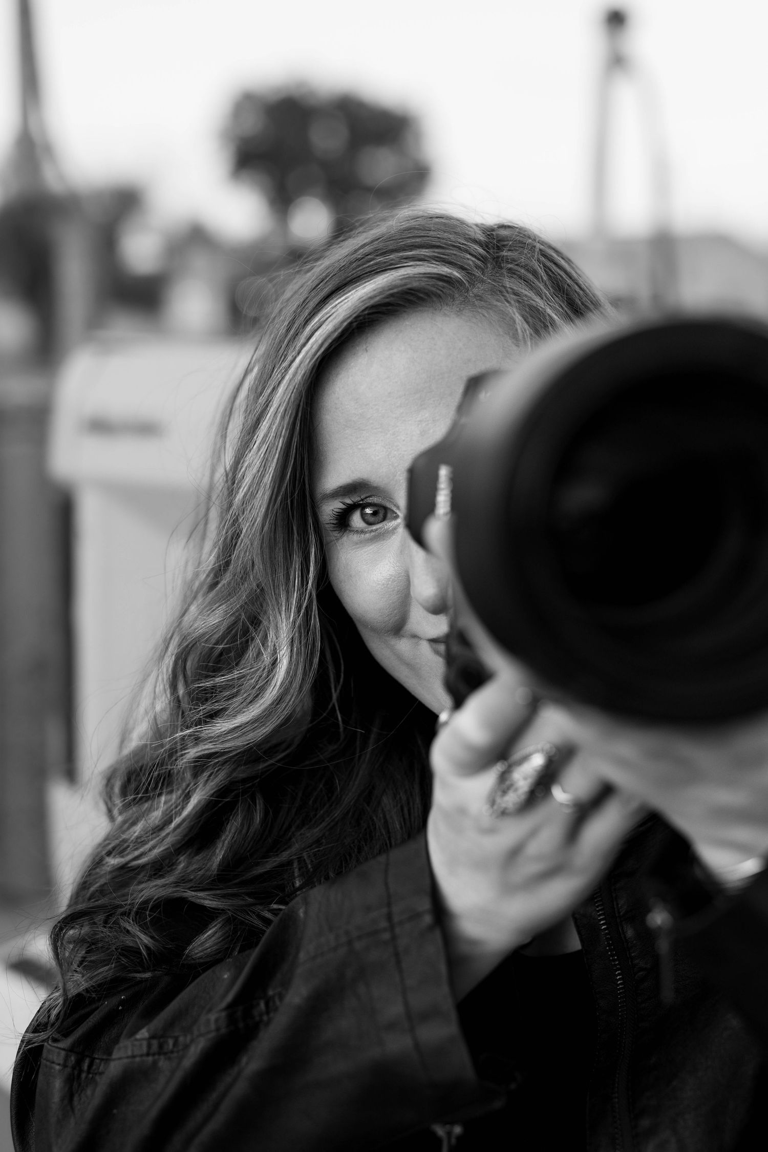 Black and white portrait of a woman with long hair holding a camera lens up to her face outdoors.