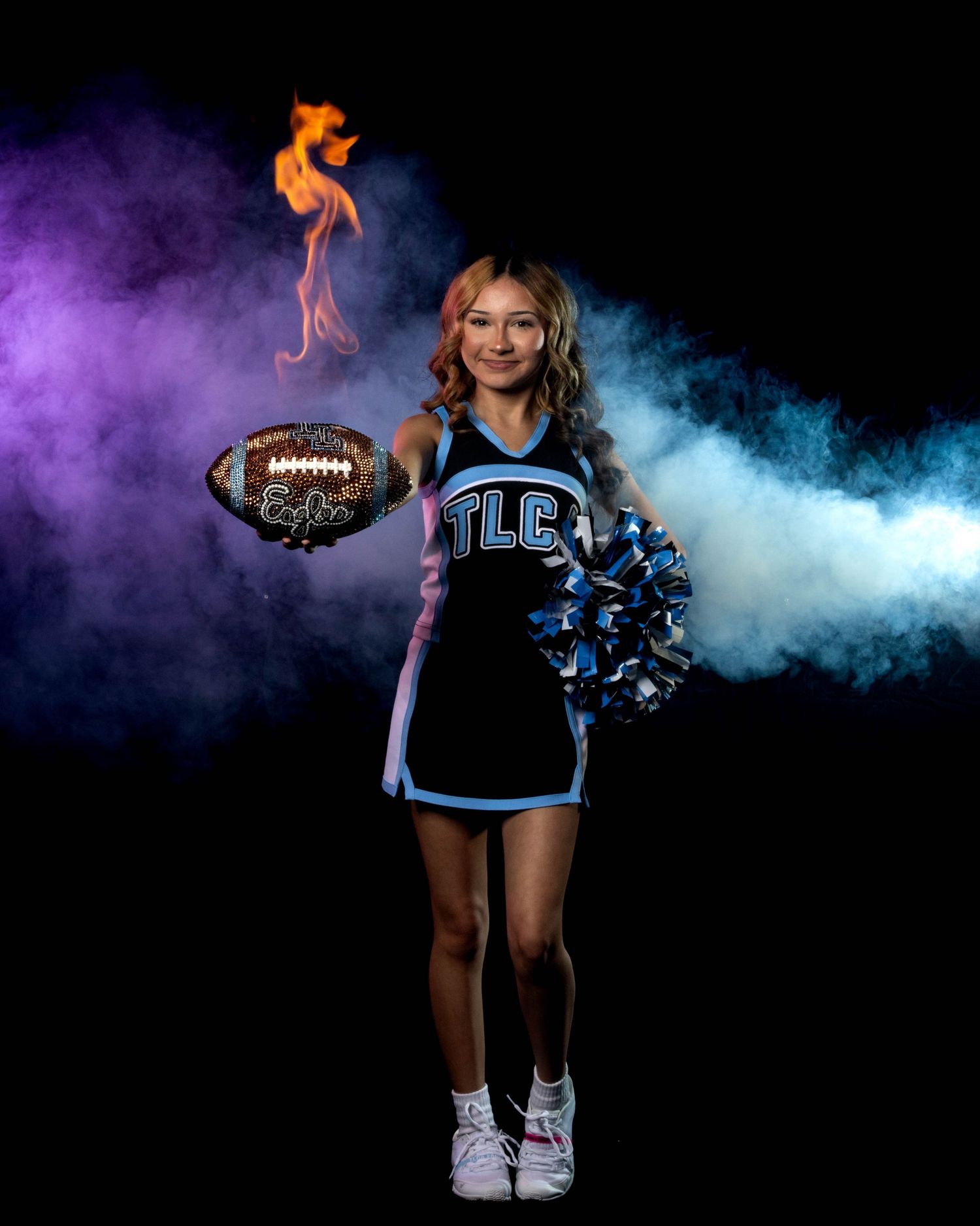 Cheerleader in TLC uniform holds a flaming football with pom-poms in a colorful smoky studio portrait.
