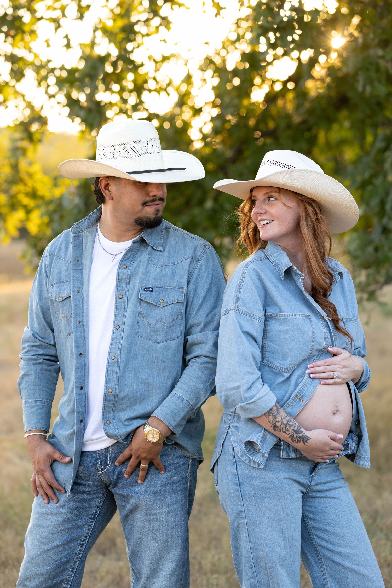 A pregnant couple in matching denim outfits and cowboy hats pose outdoors, smiling at each other in golden sunlight.