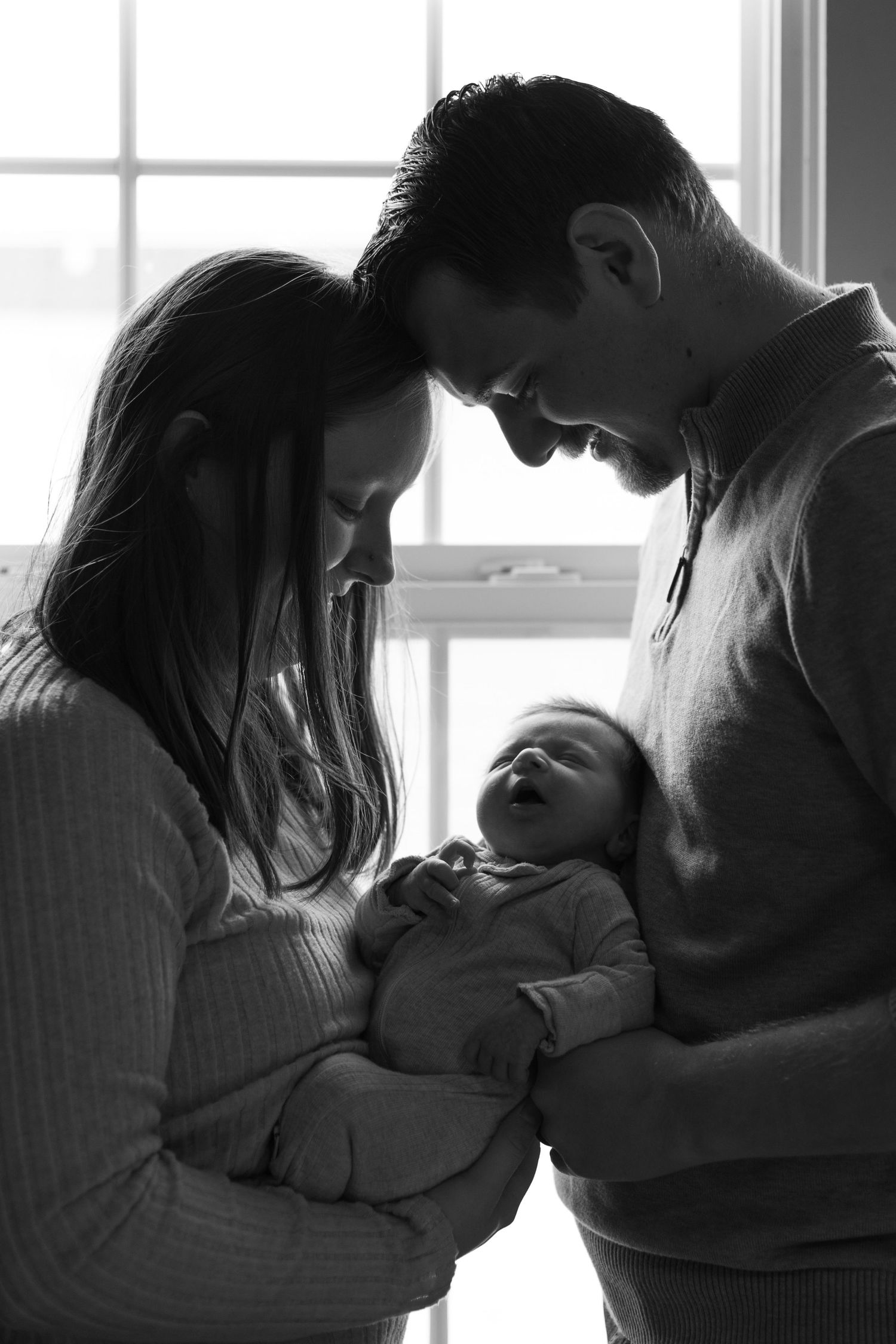 Black and white photo of parents lovingly cradling their newborn baby in front of a window.