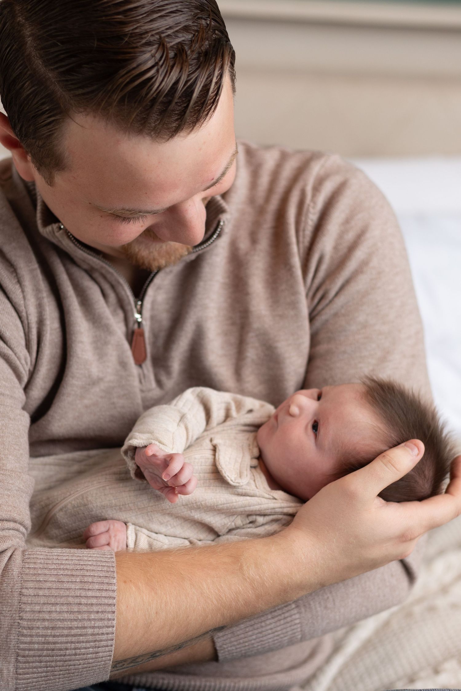 Father in beige sweater tenderly cradling his newborn baby, gazing down with love and affection.