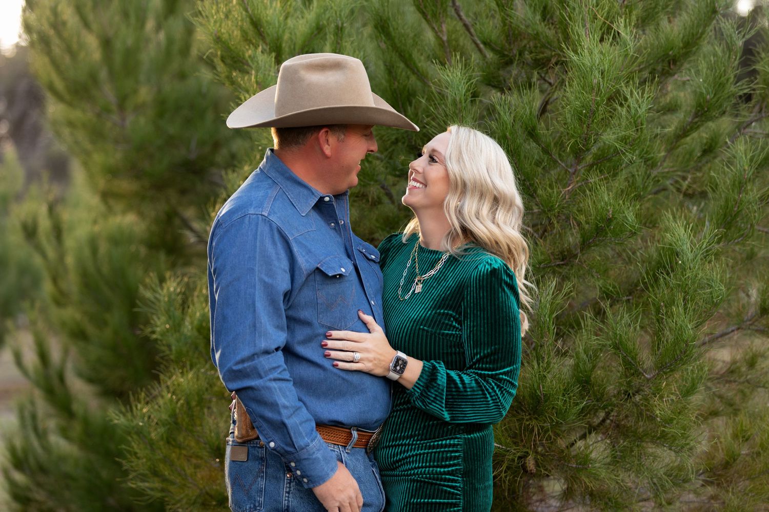 Couple in western attire smiling at each other near pine trees outdoors.