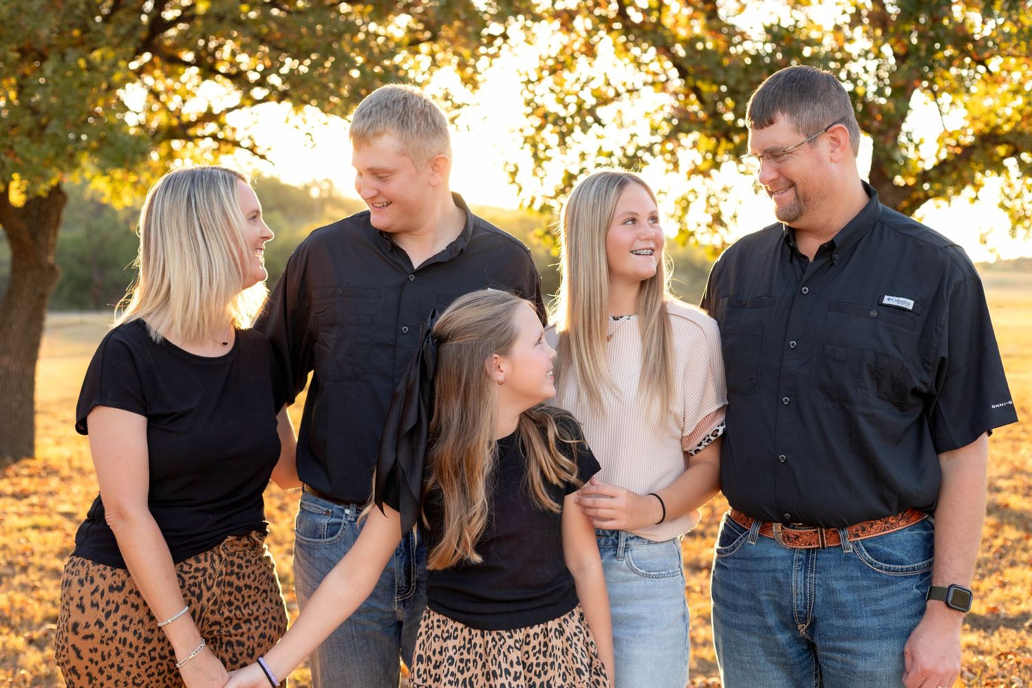 Family of five laughing together outdoors during golden hour among autumn trees.