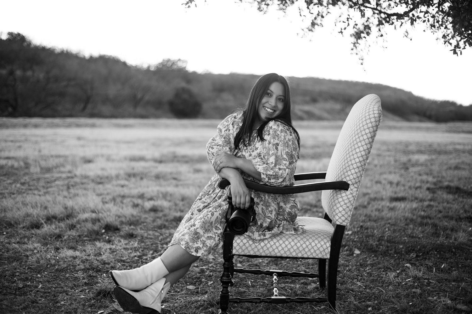 Black and white portrait of young woman in floral dress sitting on vintage chair in open field.